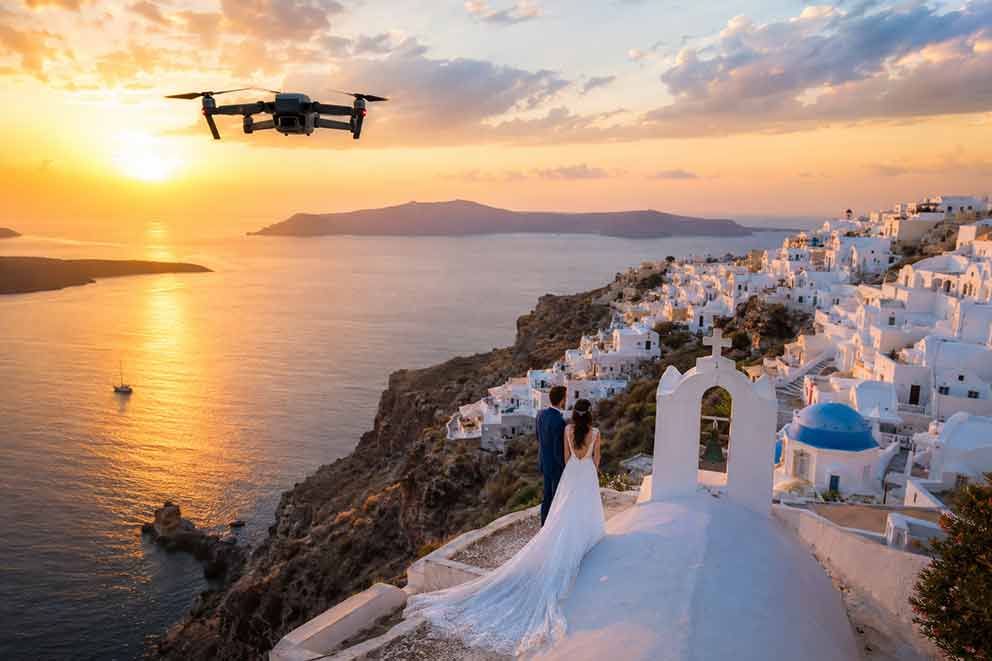 Destination wedding drone στην Ελλάδα: κινηματογραφική εναέρια λήψη ζευγαριού σε λευκό εκκλησάκι με θέα το Αιγαίο κατά το ηλιοβασίλεμα.