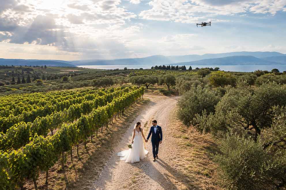 Drone στον γάμο: ζευγάρι περπατά σε αμπελώνα ενώ drone καταγράφει κινηματογραφικές εναέριες λήψεις υπό φυσικό φως.
