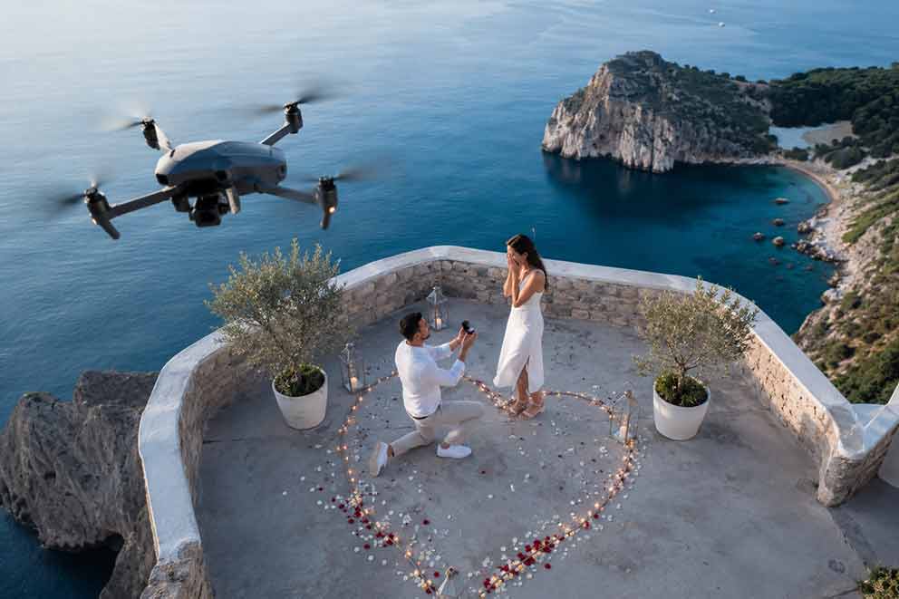 Drone για πρόταση γάμου: άνδρας γονατίζει σε παραλία κατά το ηλιοβασίλεμα ενώ drone καταγράφει κινηματογραφικά τη στιγμή από ψηλά.